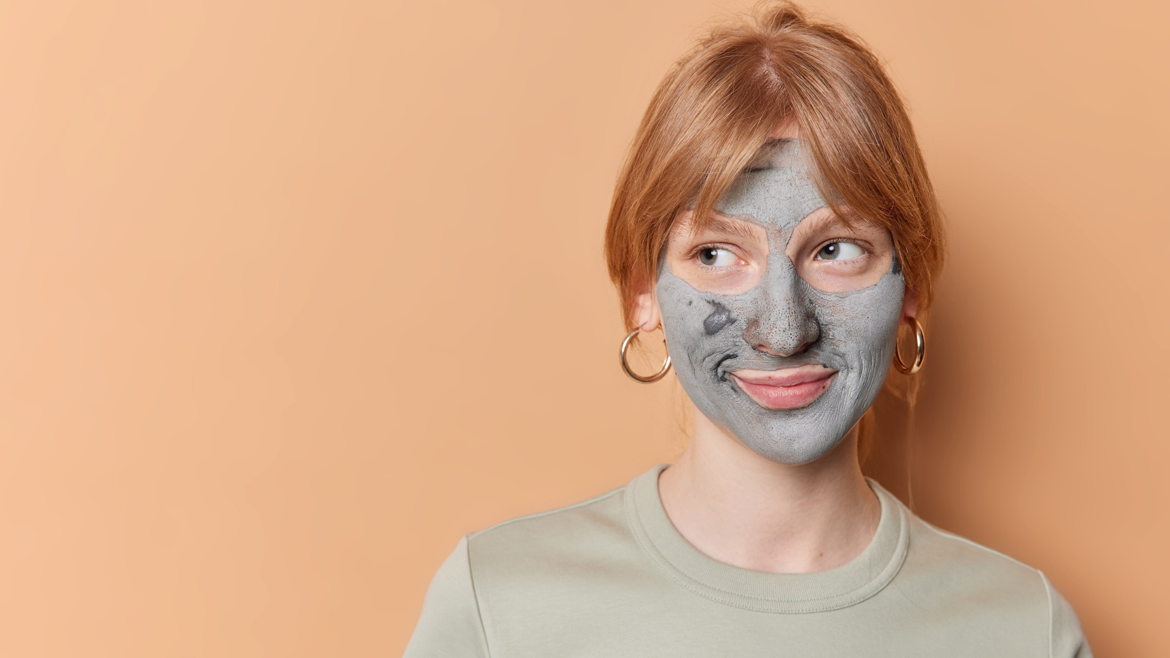 This image features a young woman with striking red hair and fair skin, prominently displaying a grey facial mask against a warm peach background
